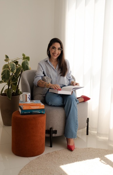 Larissa Bez lendo em uma poltrona, ao lado de uma pilha de livros