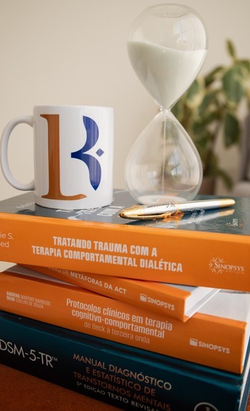 Livros técnicos de psicologia empilhados ao lado de uma ampulheta e de uma caneca com o símbolo da marca